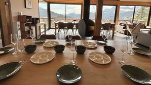 Un coin repas avec une grande table dressée pour le repas, un poêle à bois et une vue sur les montagnes par de grandes fenêtres.