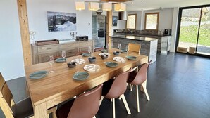 Une cuisine moderne dotée d’un îlot central, de poutres en bois au plafond et d’un coin repas avec une grande table dressée pour six personnes.