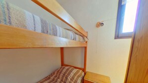 Un lit superposé avec une couverture à carreaux, une table de chevet en bois avec des tiroirs et une fenêtre avec des stores.