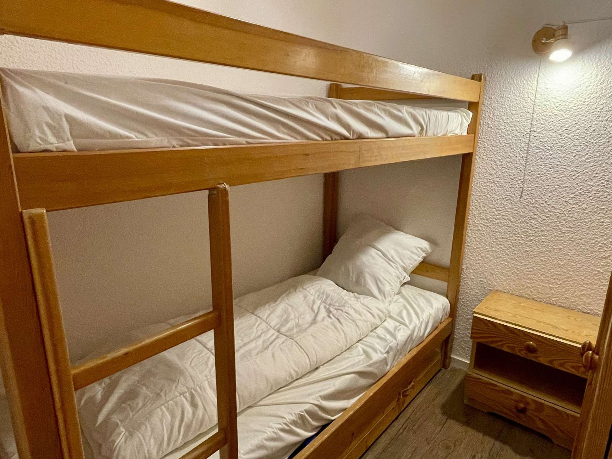A bunk bed with a wooden frame and white bedding. There is a small wooden nightstand with drawers.