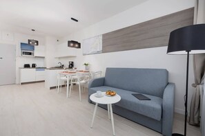 Ein modernes Wohnzimmer mit einem blauen Sofa, einem kleinen weißen Tisch und einer Lampe. Im Hintergrund befindet sich eine Küchenzeile mit weißen Schränken und einer Mikrowelle.