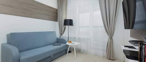 Ein modernes Wohnzimmer mit einem blauen Sofa, einem kleinen weißen Tisch, einer Stehlampe, einem Fernseher und einem Bücherregal.