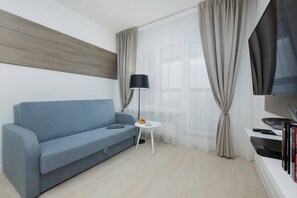 Ein modernes Wohnzimmer mit einem blauen Sofa, einem kleinen weißen Tisch, einer Stehlampe, einem Fernseher und einem Bücherregal.