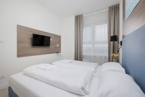 A hotel room with a bed, a TV mounted on a wooden panel, a window with curtains, and a vase with flowers.
