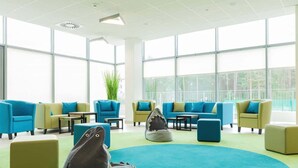 A modern lounge area with sofas and chairs arranged around a central rug, large windows providing natural light, and a plant adding greenery.