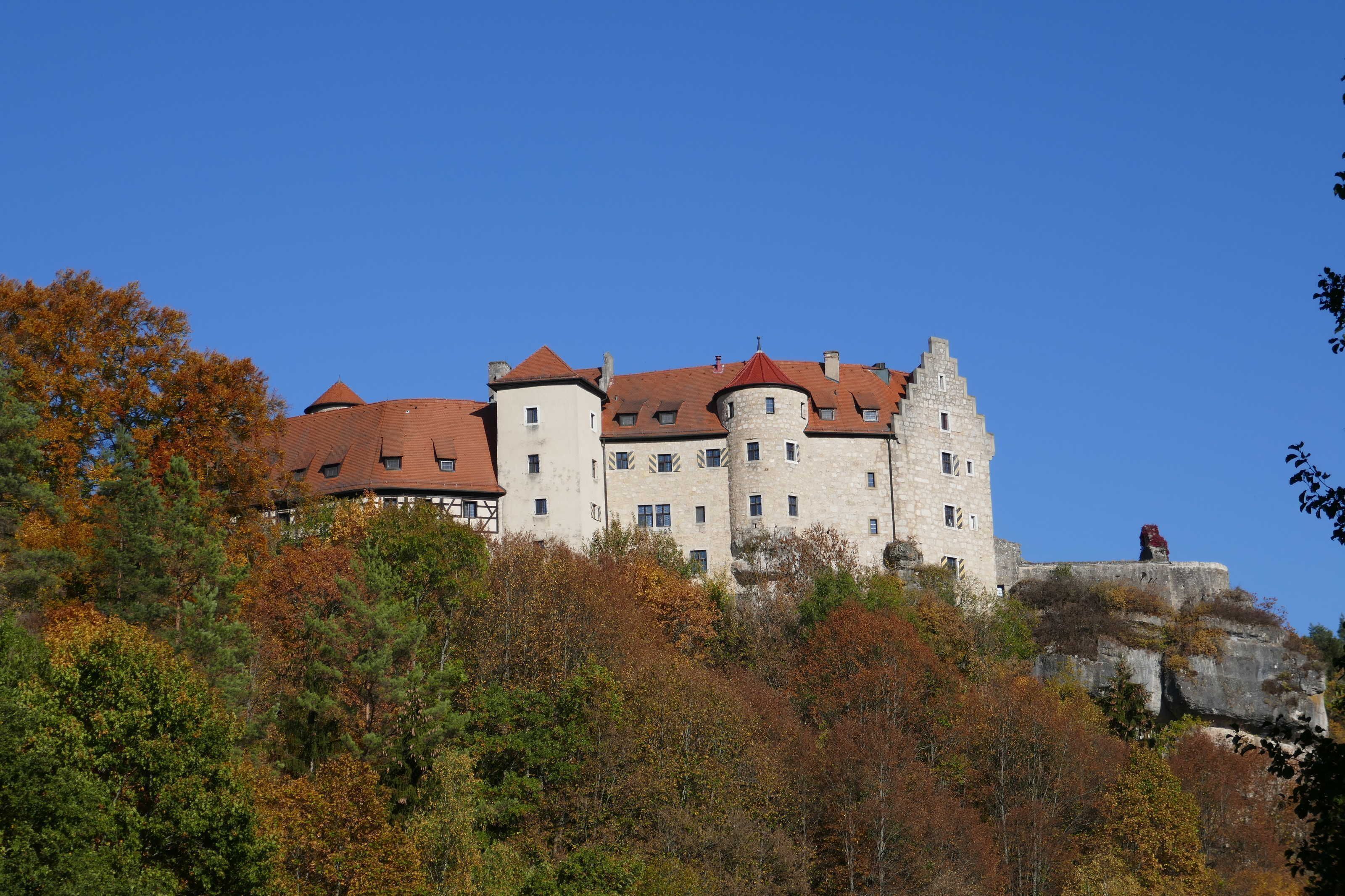 Un bâtiment qui ressemble à un château, avec de nombreuses tours et un toit rouge, entouré d’un feuillage automnal.