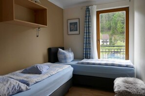 A bedroom with two beds, a window with a view of a house and trees, a wooden shelf, and a framed picture on the wall. - Limerhof Vacation Cottage - Vacation Cottage (Waischenfeld)