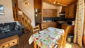 A cozy room with a sofa, a dining table set for four, wooden cabinets, a staircase, and a kitchen area. - Cozy apartment in Les Gets with balcony and parking (Les Gets)