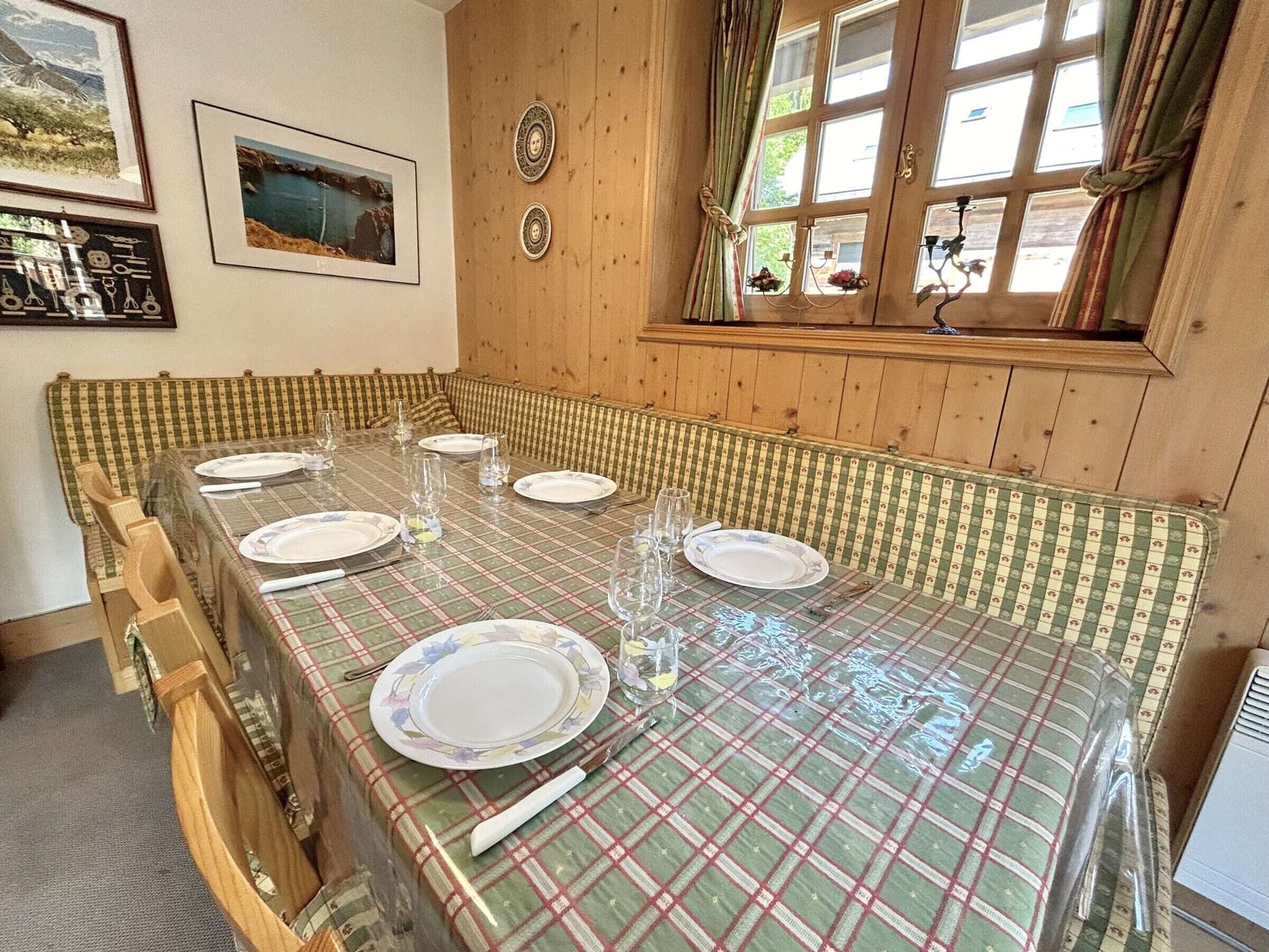 A dining room with a long table set for four, wooden paneled walls, and two framed pictures on the wall.