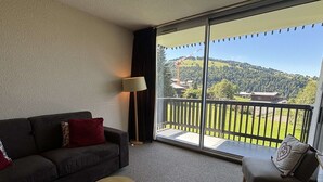 A room with a sofa, a chair, a small round table, and a large window with a view of a green landscape and a building. - 2-room apartment, pets allowed, parking in Notre-Dame-de-Bellecombe (Notre-Dame-de-Bellecombe)