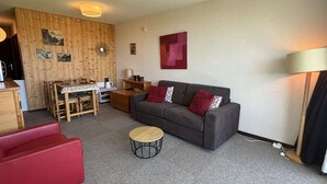 A cozy living room with a grey sofa, red armchair, and a small round coffee table. A wooden dining table with chairs is against the wall, and a lamp is on the floor. - 2-room apartment, pets allowed, parking in Notre-Dame-de-Bellecombe (Notre-Dame-de-Bellecombe)