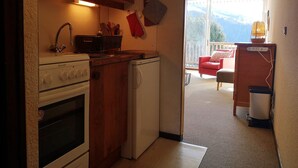 A compact kitchen with wooden cabinets, a stove, and a refrigerator. A red chair and a small table are visible in the adjacent living area, which has a balcony with a view of the mountains. - 2-room apartment, pets allowed, parking in Notre-Dame-de-Bellecombe (Notre-Dame-de-Bellecombe)