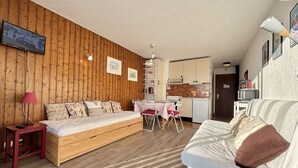 A cozy room with a wooden wall, a bed, a sofa, a dining table with chairs, a kitchen area, and a TV mounted on the wall.