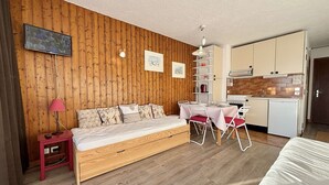 A cozy room with a wooden wall, a bed, a dining table with chairs, a kitchen area, and a TV mounted on the wall.