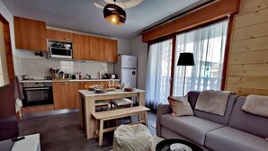 A modern living room with a grey sofa, a wooden dining table, and a kitchen area with wooden cabinets and a built-in oven. - Renovated apartment for 4, near center, parking (Les Gets)