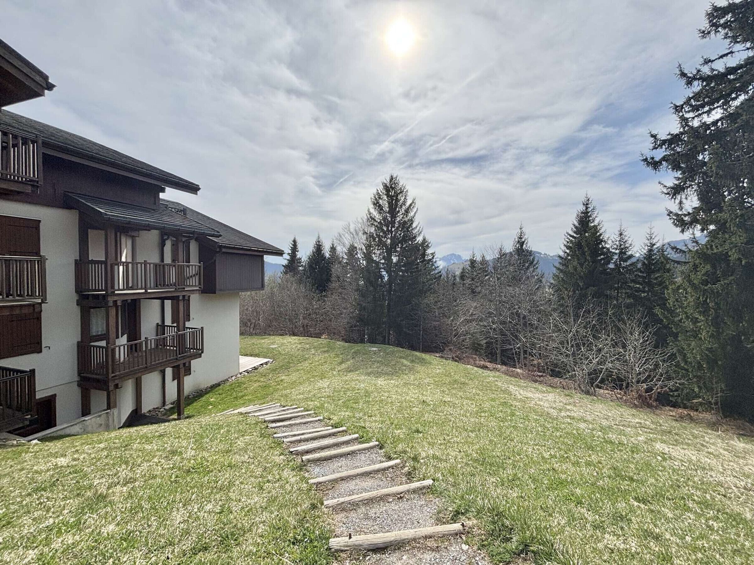 A modern two-story building with a balcony, a grassy slope leading to a wooden staircase, and a forested backdrop.