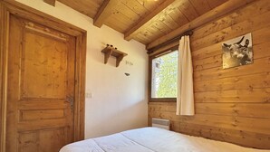 Ein Zimmer mit Holzboden, einem Bett, einer Tür, einem Fenster mit Vorhängen und einem an der Wand befestigten Regal.