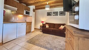 Eine kompakte Küche mit Holzausbau, einer Mikrowelle, einem Herd und einem Geschirrspüler. Im Wohnbereich befindet sich ein braunes Sofa und ein kleiner Tisch mit einer Lampe.