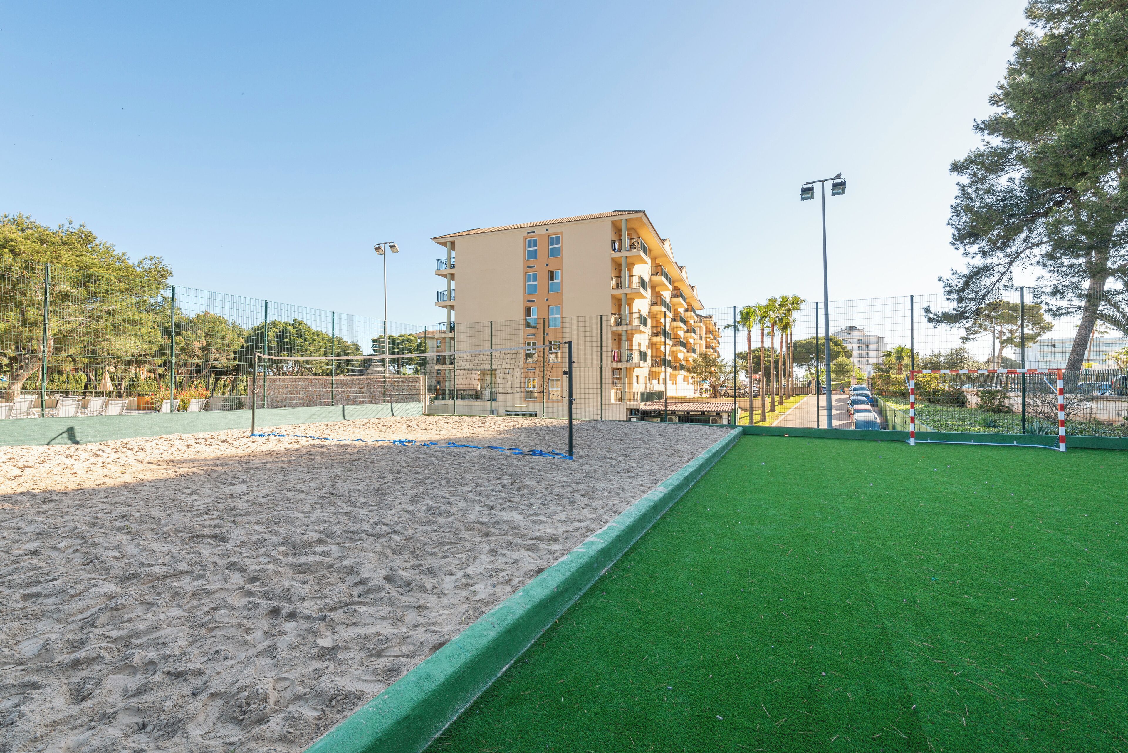 A sports court with sand and artificial turf, surrounded by a fence and a building with multiple balconies.