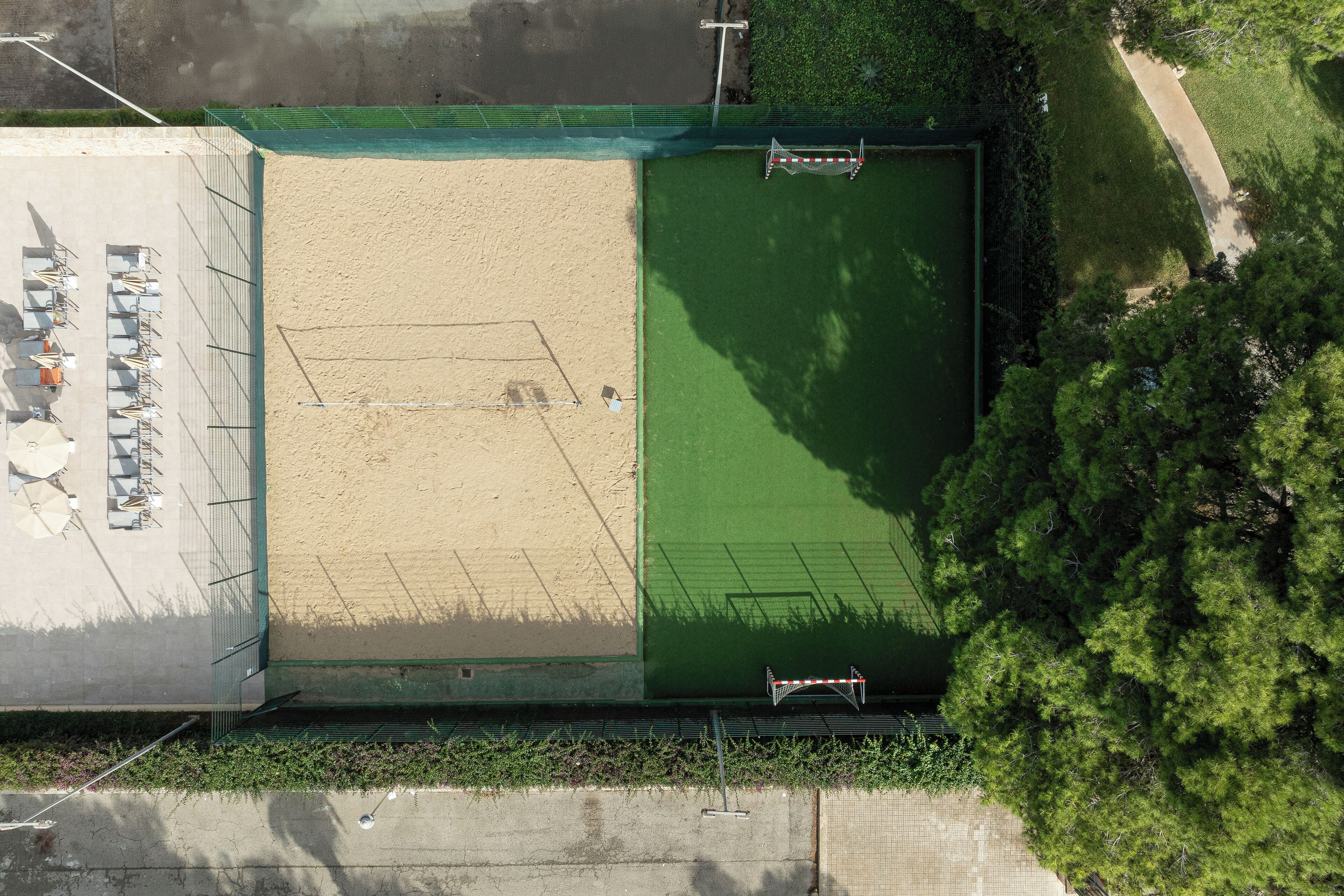 Vista aérea de una cancha deportiva con zona de arena y zona de césped verde, rodeada de árboles y una valla.