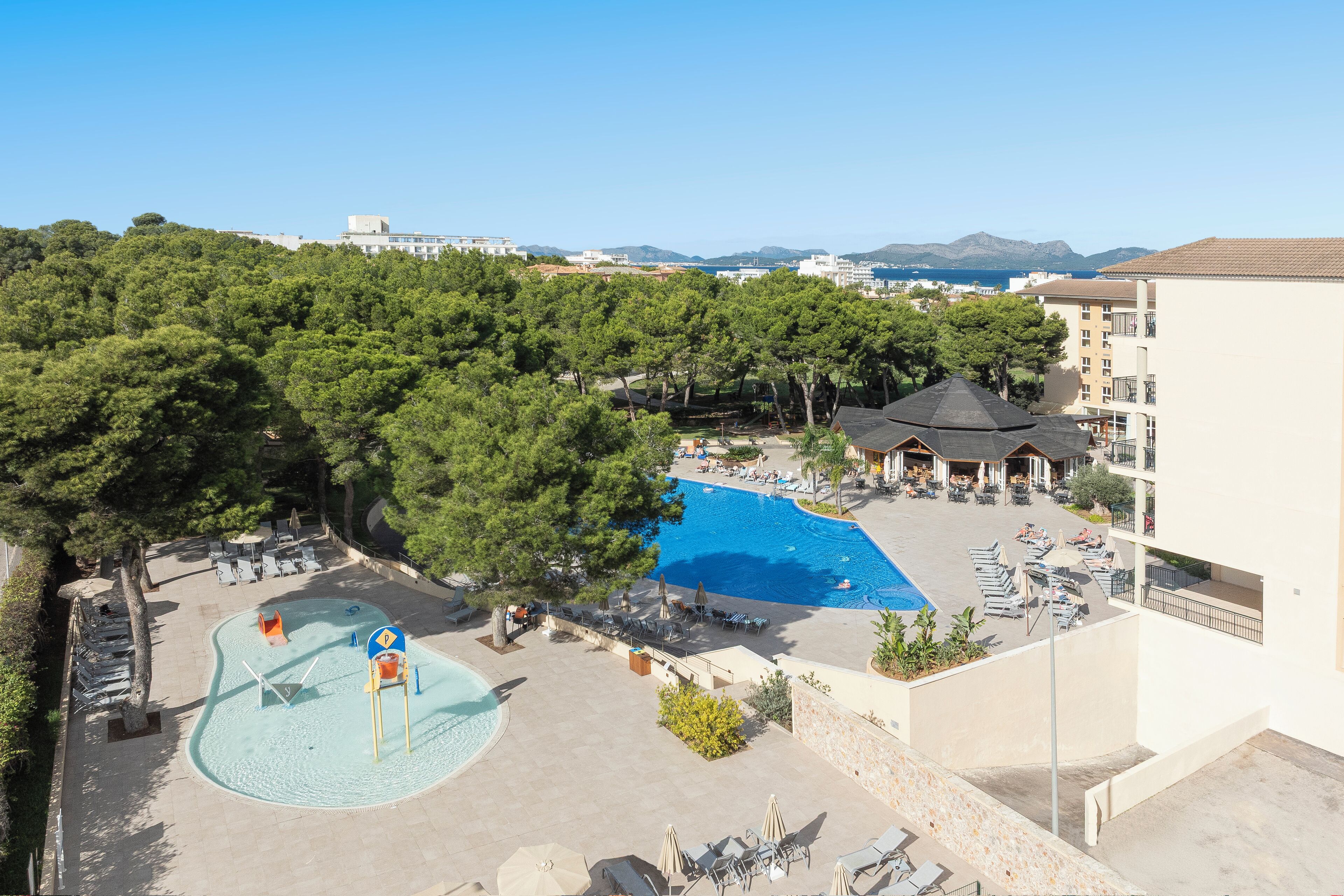 Un resort con una gran piscina rodeada de sillas de descanso y sombrillas, un área de juegos para niños y vistas al mar y a las montañas.