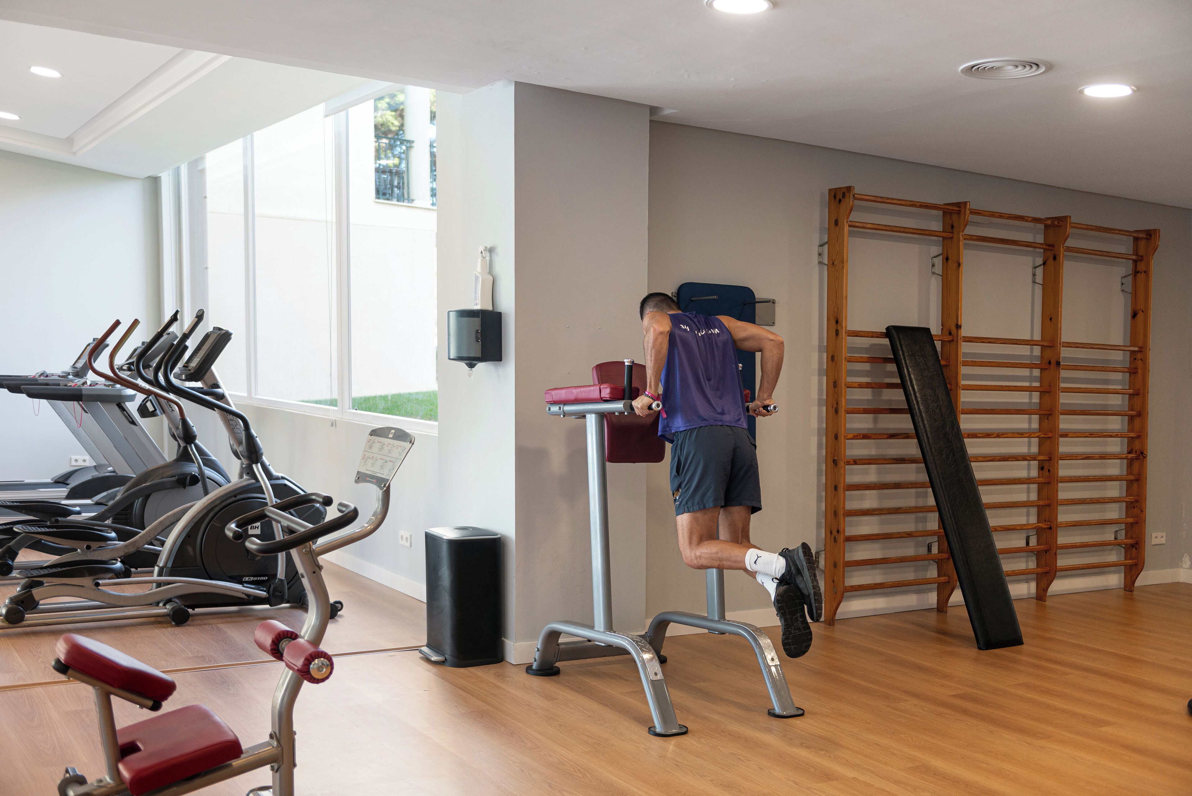 Un gimnasio con cintas de correr, una plataforma elevada y una pared de escalar de madera.