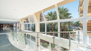 A spacious indoor area with a curved glass railing, a view of palm trees, and a clear sky. - Aparthotel Can Picafort Palace (Santa Margalida)