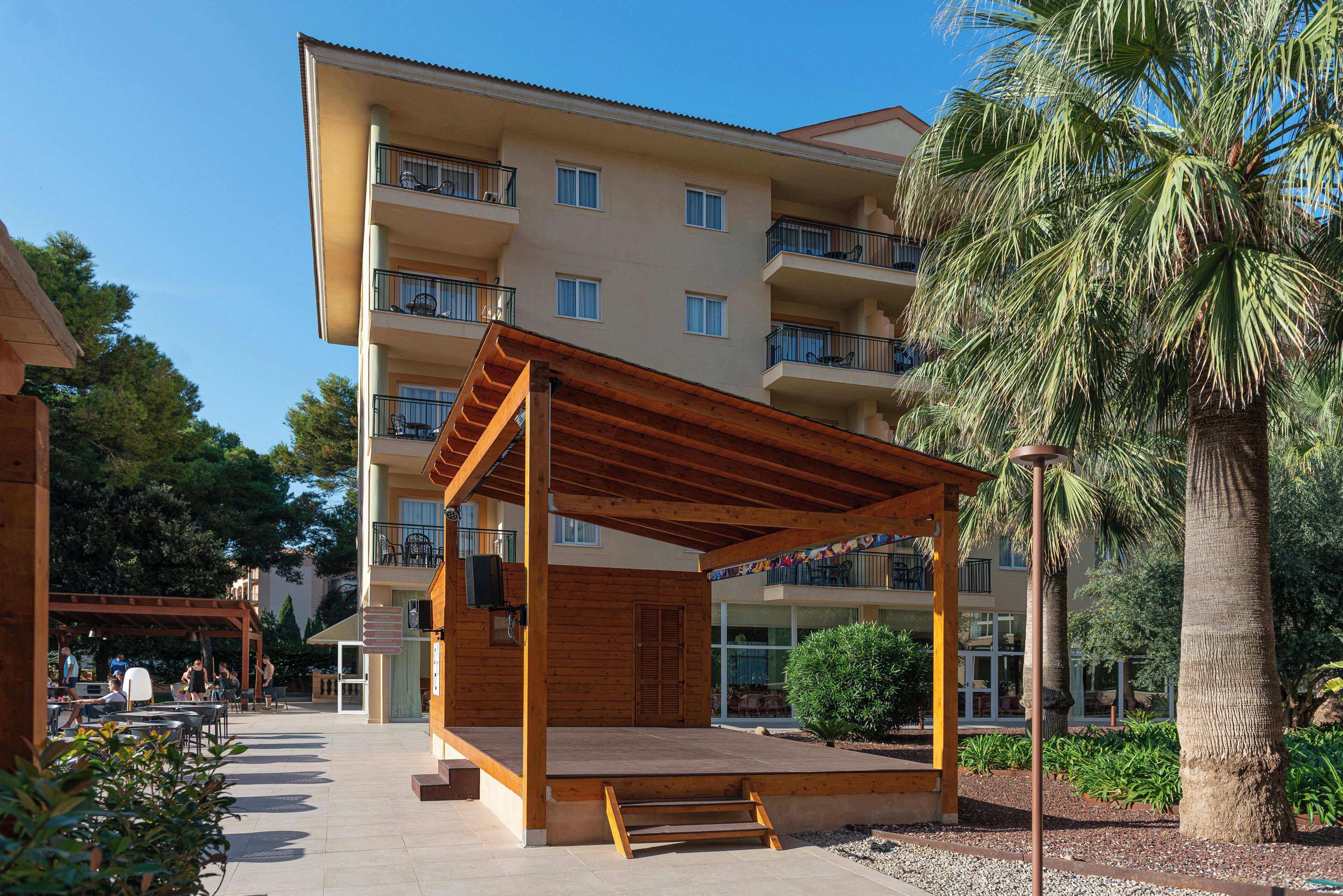 Un hotel de varios pisos con balcones, un área de estar exterior cubierta y palmeras.