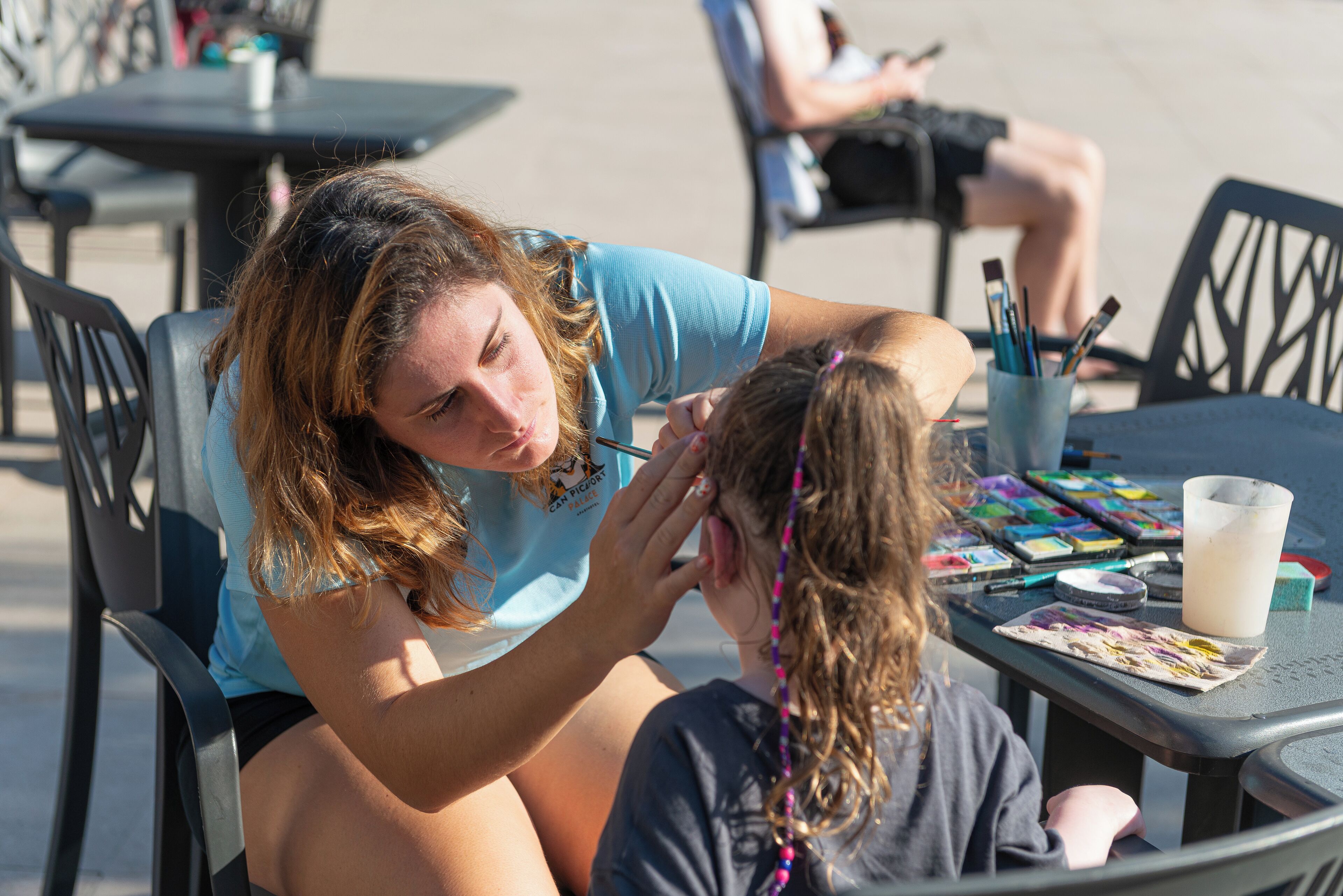 Une personne est en train de maquiller le visage d’un enfant à une table installée à l’extérieur, en utilisant du matériel de peinture.