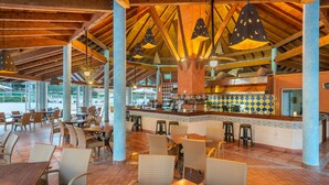 A spacious indoor dining area with high ceilings, wooden beams, and hanging pendant lights. Tables and chairs are arranged for dining, and a bar area is visible in the background. - Aparthotel Can Picafort Palace (Santa Margalida)