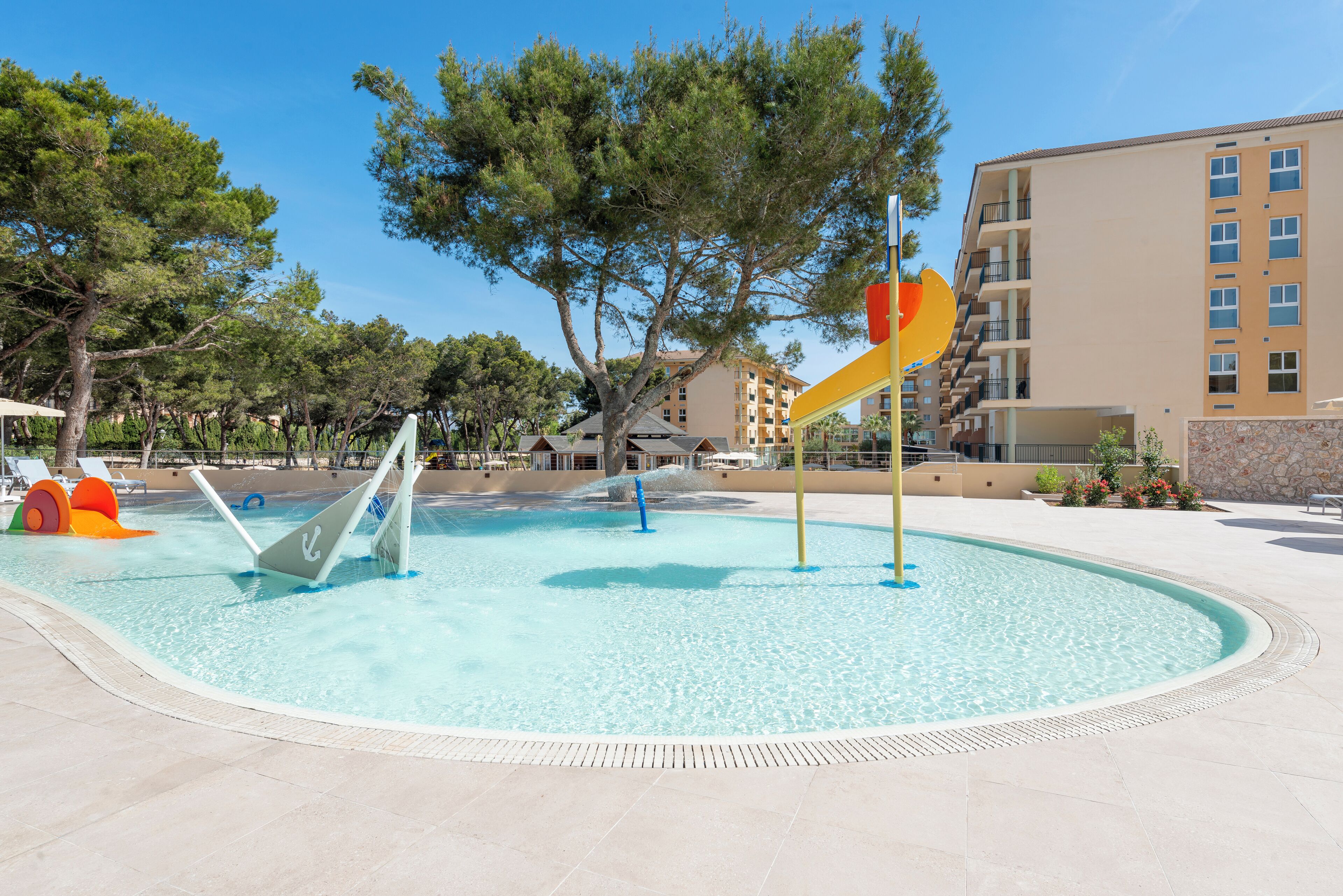 A swimming pool with slides and water features, surrounded by trees and a building with balconies.