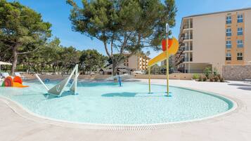 A swimming pool with slides and water features, surrounded by trees and a building with balconies.