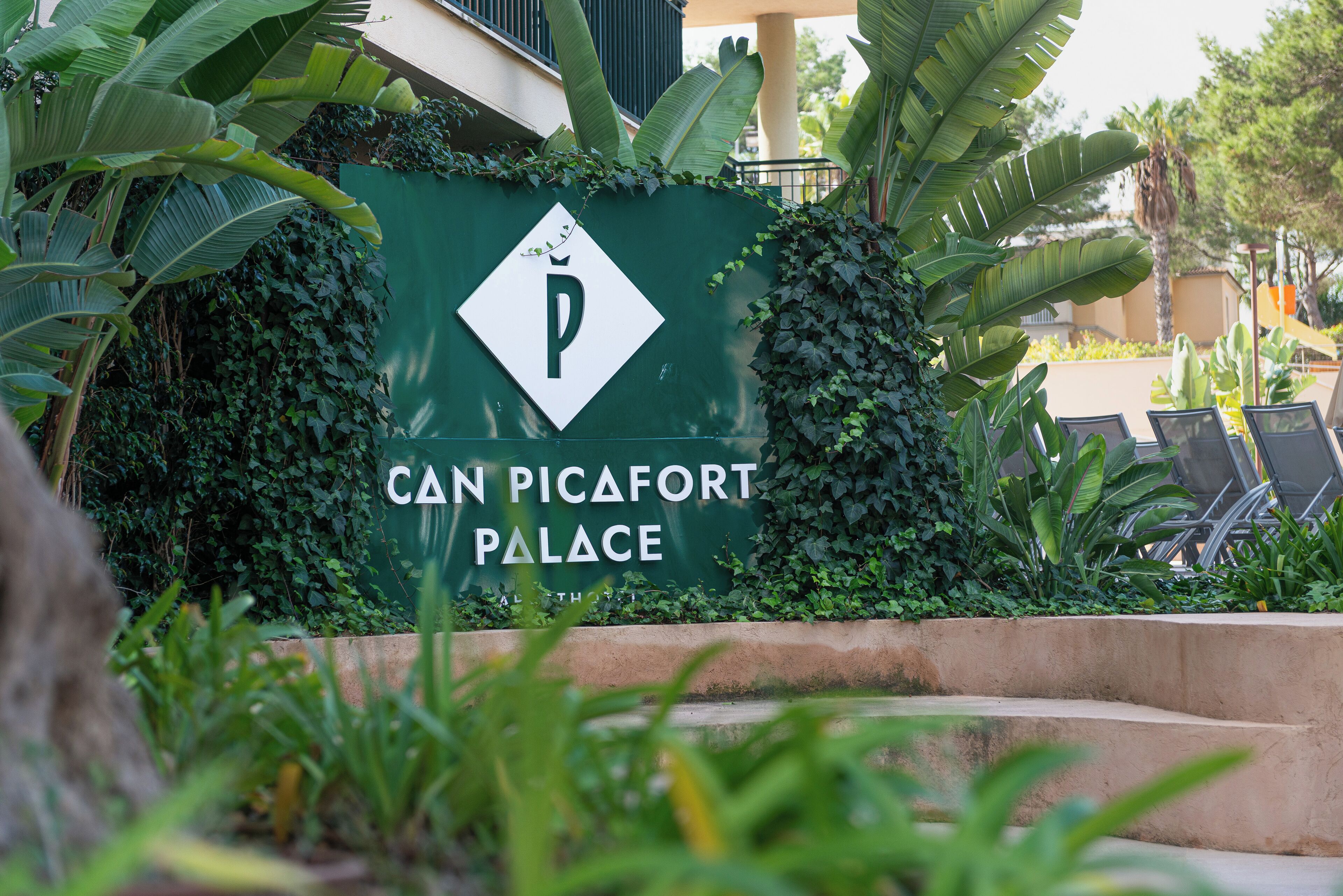 Señal del Palacio Can Picafort con símbolo de estacionamiento, rodeada de vegetación exuberante.