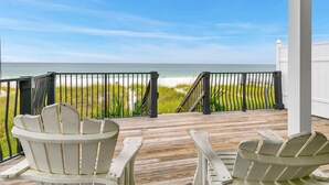 A wooden deck with two Adirondack chairs facing a beach with a metal railing.