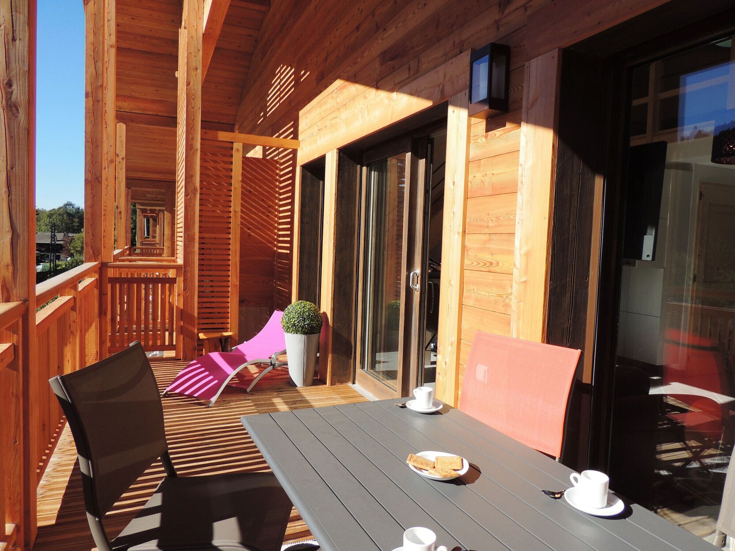Een houten terras met een tafel, stoelen, een plant in pot en een schuifdeur met glas.