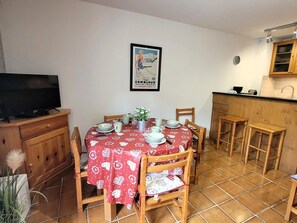A dining area with a red tablecloth, wooden chairs, a television on a cabinet, and a kitchen area with a wooden counter and cabinets.