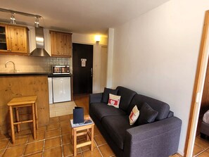 A compact living space with a kitchenette, a dark grey sofa, a small wooden stool, and a tiled floor.
