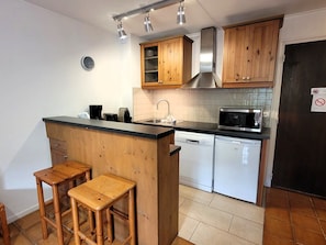 A compact kitchen with wooden cabinets, a microwave, a dishwasher, and two stools.