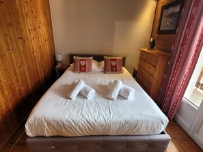 A hotel room with a bed, two heart-patterned pillows, a wooden dresser, and a nightstand with a lamp.
