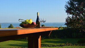A table with a bottle and glasses, a basket of fruit, and a view of the sea.