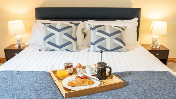 Una cama bien hecha con funda de almohada azul y blanca a cuadros, una bandeja de madera con alimentos para el desayuno y lámparas de noche en las mesitas.