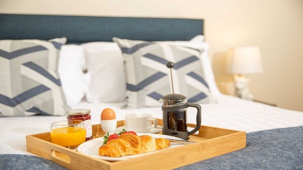 A breakfast tray on a bed with a coffee maker, croissant, orange juice, and jam.