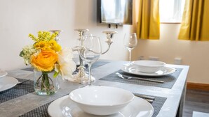 Una mesa de comedor dispuesta con platos, tazones y cubiertos blancos, un florero con flores amarillas y una copa de vino.