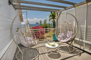A balcony with two hanging chairs, a small table with drinks, and a view of the sea and a distant island.