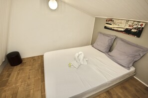 Una cama individual con colchón blanco y dos almohadas, un cesto de basura redondo negro, y un cartel colgado en la pared.