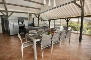 Una cocina moderna con electrodomésticos de acero inoxidable, un comedor con una mesa dispuesta para cuatro personas, y un patio cubierto con vistas a zonas verdes.
