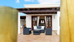 An outdoor patio area with wicker furniture, a tiled floor, and a white building in the background. - Casa La Goleta, Volcán y Mar (Playa Blanca)