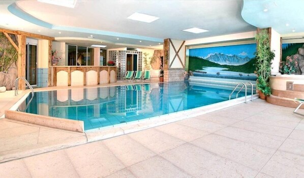 Una piscina interior con vistas a montañas en la pared, un área de descanso con sillas y una escalera que conduce a un nivel superior.