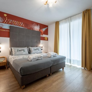 Une chambre à coucher moderne avec un grand lit, une tête de lit et une vue sur l’extérieur à travers des rideaux transparents.