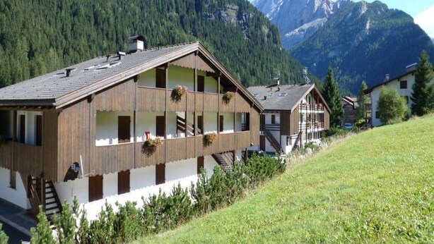 Ein Chalet mit geneigtem Dach, Holzfassade und mehreren Balkonen mit Blumenkästen. Es liegt in einer Bergregion mit freiem Blick auf die umliegenden Gipfel.